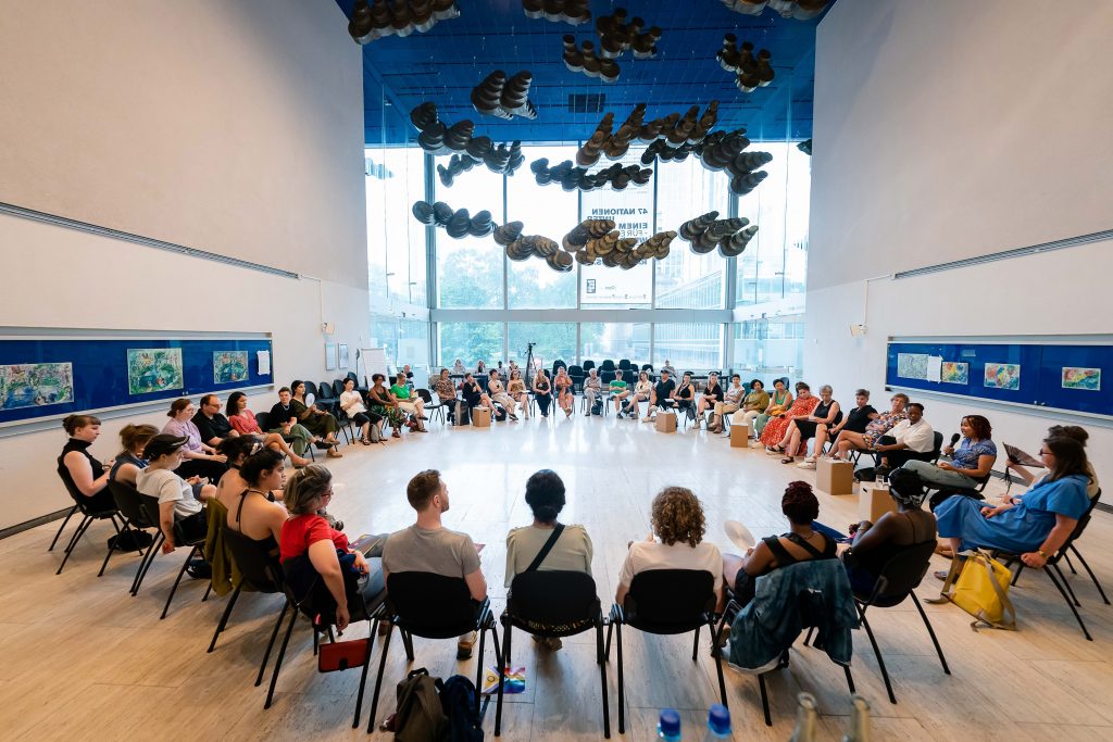 in einem großen Saal sitzen ca. 100 Menschen in einem riesigen Stuhlkreis zusammen.