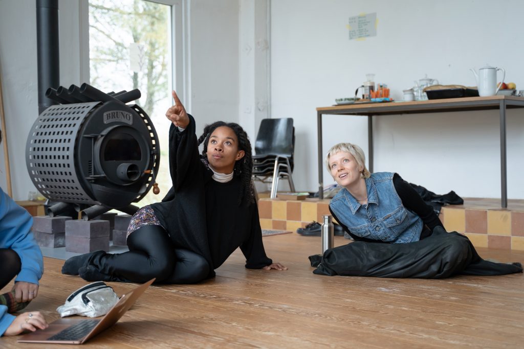 2 people sit on the floor and face the same direction. One of them excitedly points at something out of the picture.