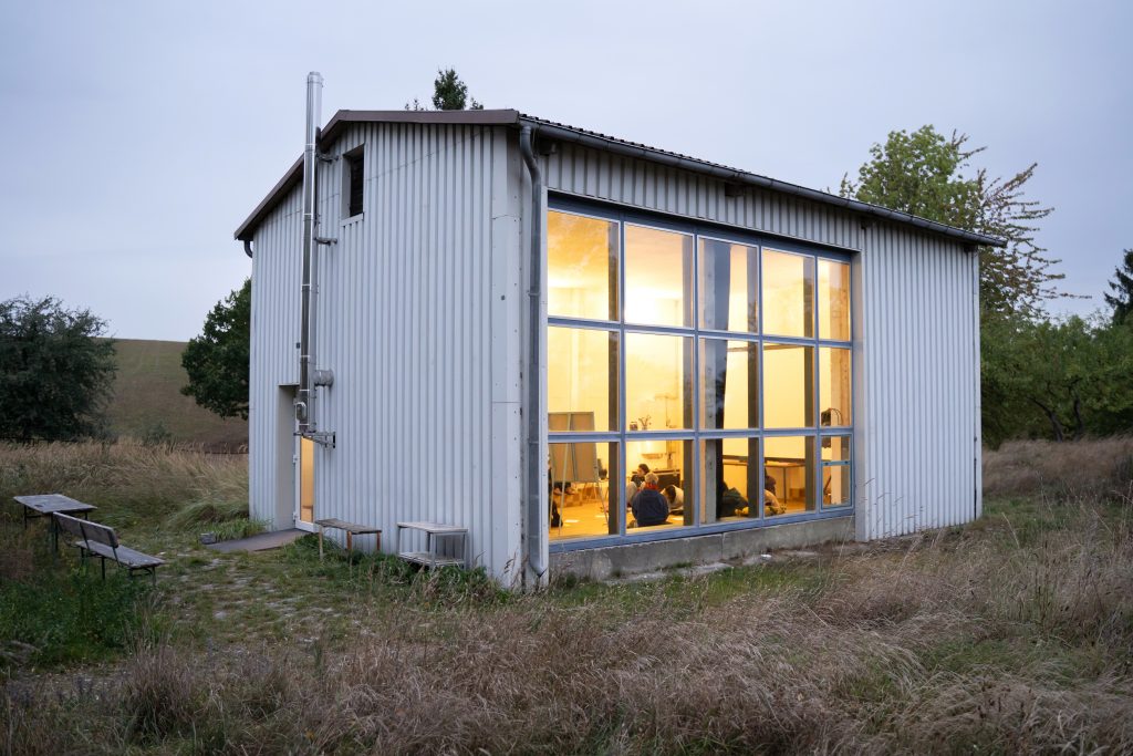 Ein kleines, weißes Häuschen steht auf einer Wiese. Es ist dämmerig - durch das große Fenster strahlt warmes, gelbes Licht nach draußen.